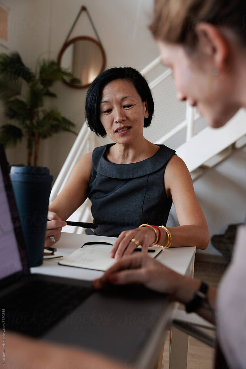 An woman in a sleeveless dark grey blouse sits at a table, looking thoughtfully across at a colleague while holding a pen over an open notebook. In the foreground, the blurred arm and hand of another person are visible typing on a laptop.