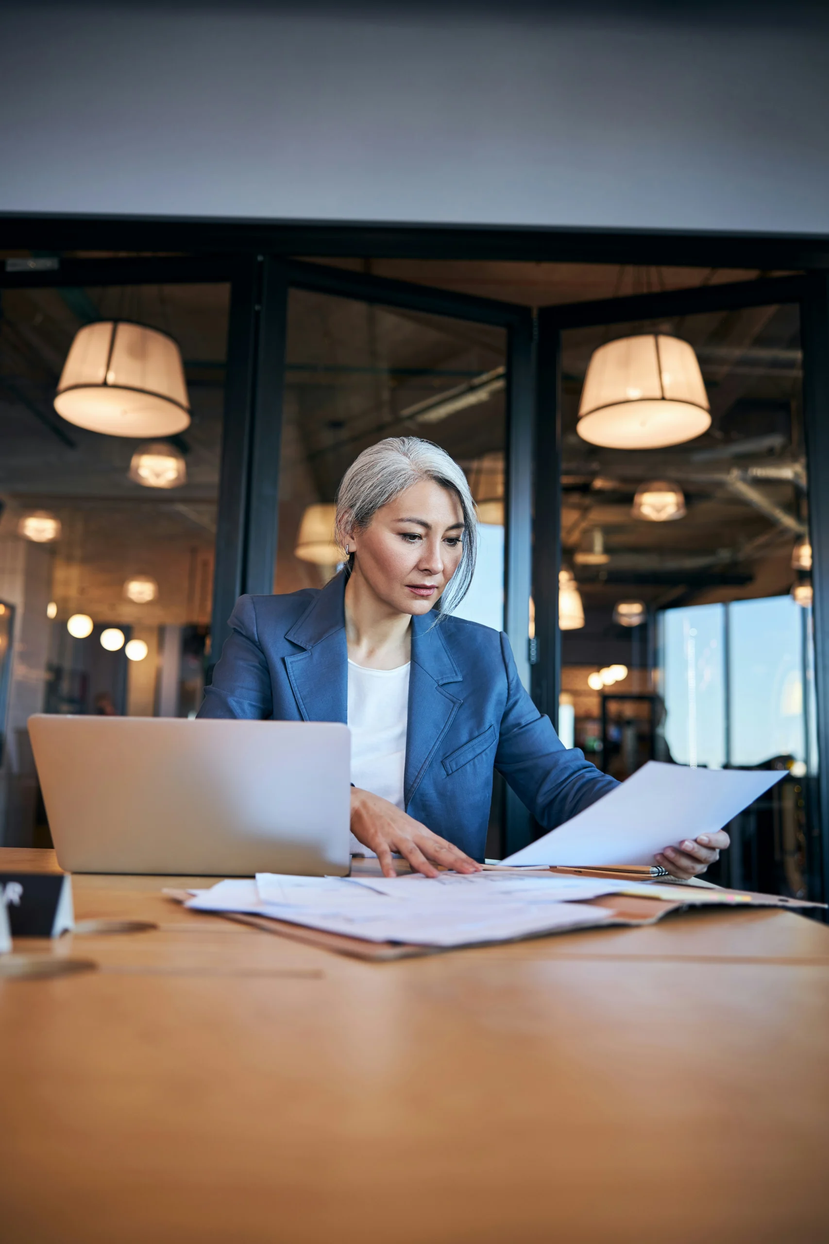 A mature woman with grey hair, wearing a blue blazer and white top, sits at a large wooden desk in a modern office with large hanging lamp fixtures. She is examining paper documents with an open laptop nearby.