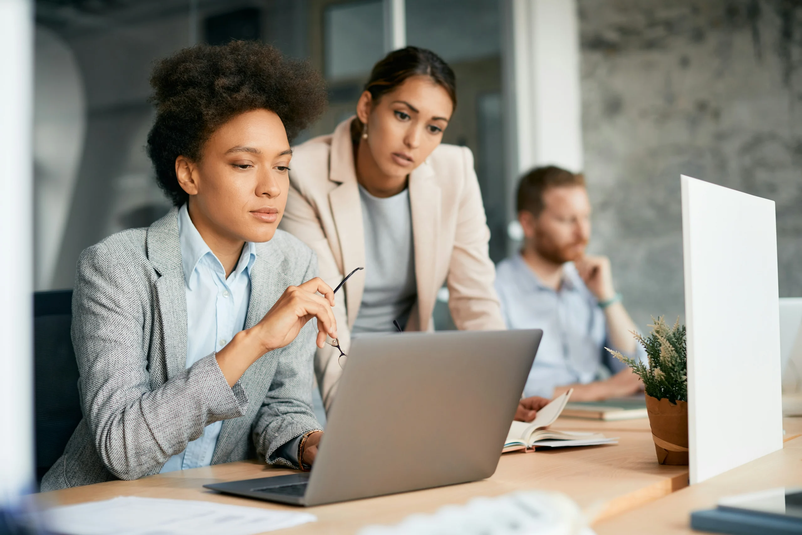 A businesswoman in a grey blazer reviews information on a laptop at a shared desk while a colleague in a beige blazer leans over to look closely at the screen. A third colleague works in the blurred background.